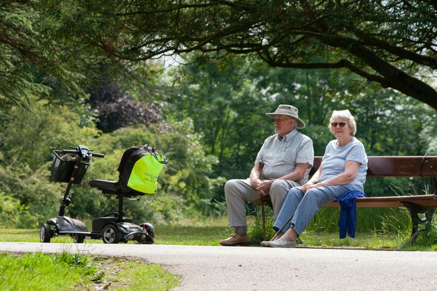 elderly man and woman sitting on a park bench with a mobility scooter next to them