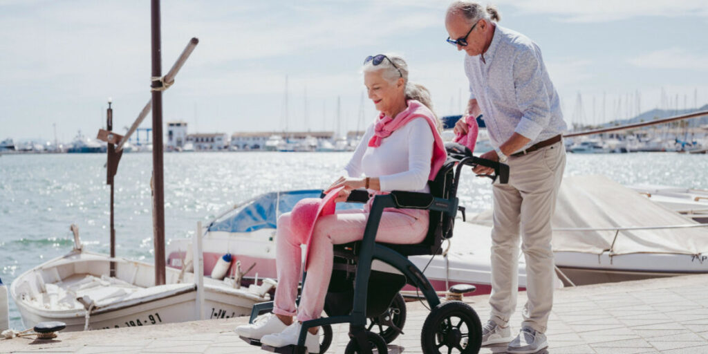lady sitting in a wheelchair holding her pink hat, with a man behind putting something in a bag on the back of the scooter. On the promenade of a quay with glistening water and boats behind.