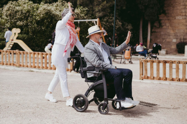 man in a a mobility scooter being pushed by a lady, both have their arms raised in greeting to someone
