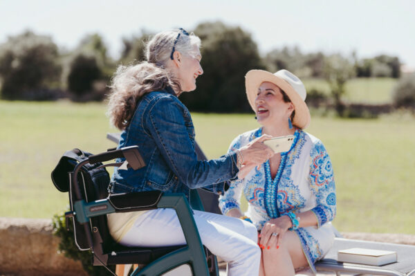 lady in a mobiltiy aid laughing with her friend who is sitting on a bench in a green field