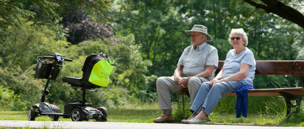 Man and woman sitting on a bench in a park with a mobility scooter