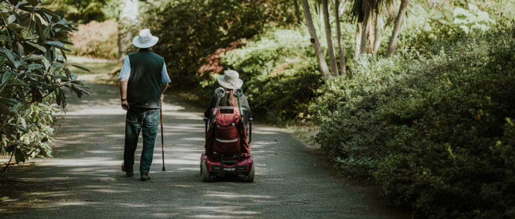 couple on a path in a park - a man walking with a stick and a lady in a mobility scooter