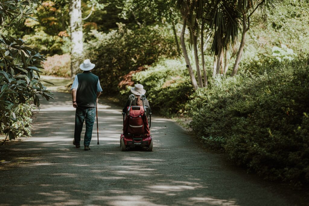 Lady on mobility scooter alongside a man with a walking stick on a path in a leafy park