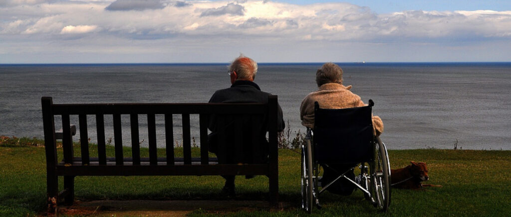 A man on a bench and a woman in a wheelchair looking out at the sea