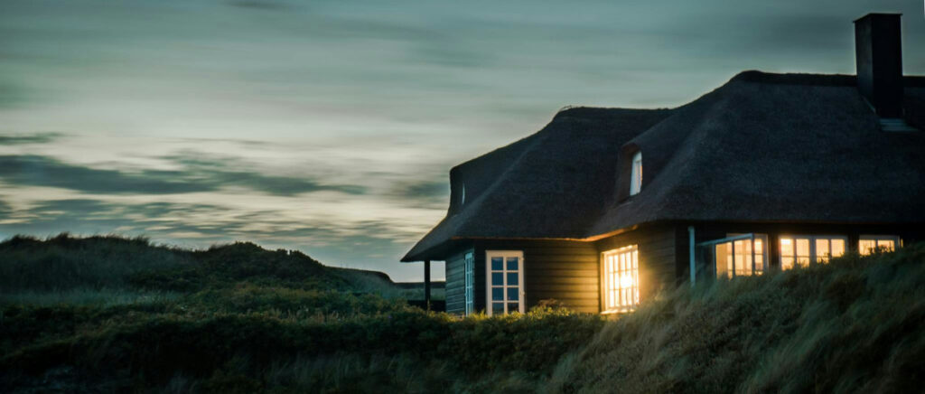 A house with a thatched roof surrounded by grass. It is dusk and there are lights on in the house.