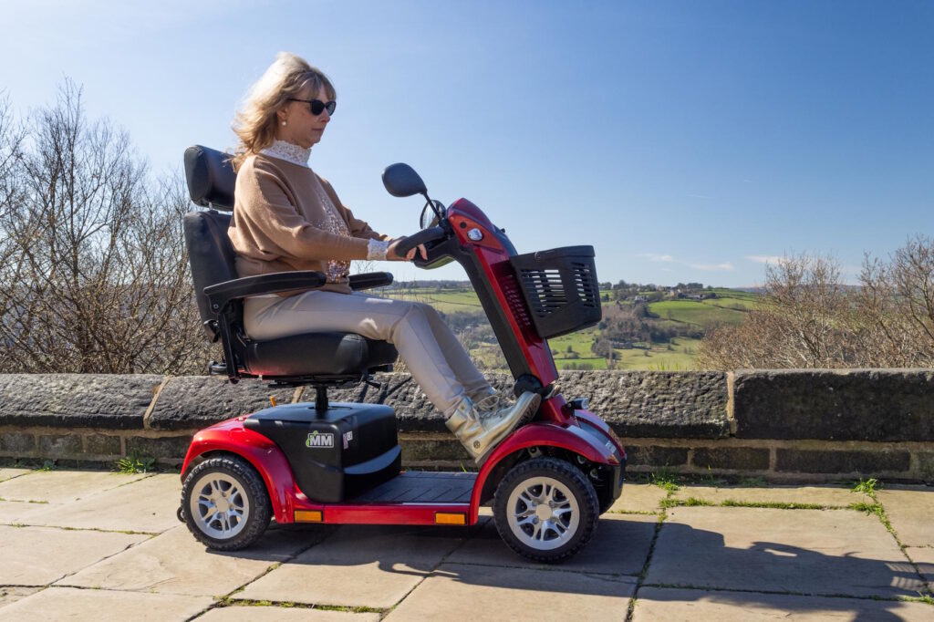 a woman riding a red mobility scooter along a pavement with blue sky and green fields in the background